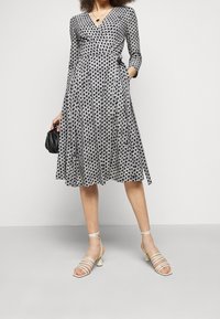 Schwarz-weißer bedruckter Wickelkleid mit langen Ärmeln, ausgestelltem Rock und V-Ausschnitt. Kombiniert mit weißen Riemchensandalen und einer kleinen schwarzen Handtasche.