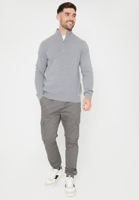 Grijze zip-neksweater met lange mouwen, gedragen met grijze cargobroeken. Witte sneakers met zwarte accenten. Eenvoudig en functioneel ontwerp.