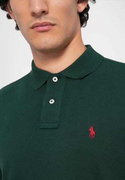 Mörkgrön polo skorta i strukturerad tyg, med spettskragen, två knappar och en liten röd broderad logotyp.