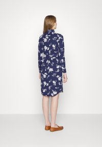 Mörkblommig klänning med långa ärmar och hög krage, med vita blommönster och en böljande fåll, i kombination med bruna platta skor.