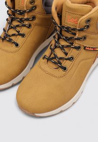 Zapatos sintéticos en tono marrón claro con una textura suave, cordones negros y detalle de logo. Cuentan con un cuello acolchado y una suela de goma blanca.