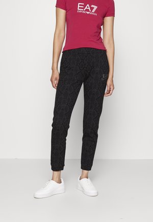 Persoon draagt een rood EA7 Emporio Armani T-shirt, zwarte joggingbroek met patroon en witte sneakers, staand tegen een effen witte achtergrond.
