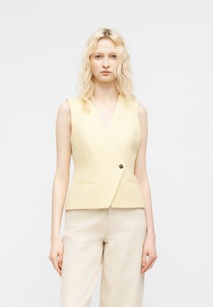 Hullámos szőke hajú nő világos sárga ujjatlan mellényt visel egy gombbal és krémszínű, texturált nadrágot egyszínű háttér előtt.