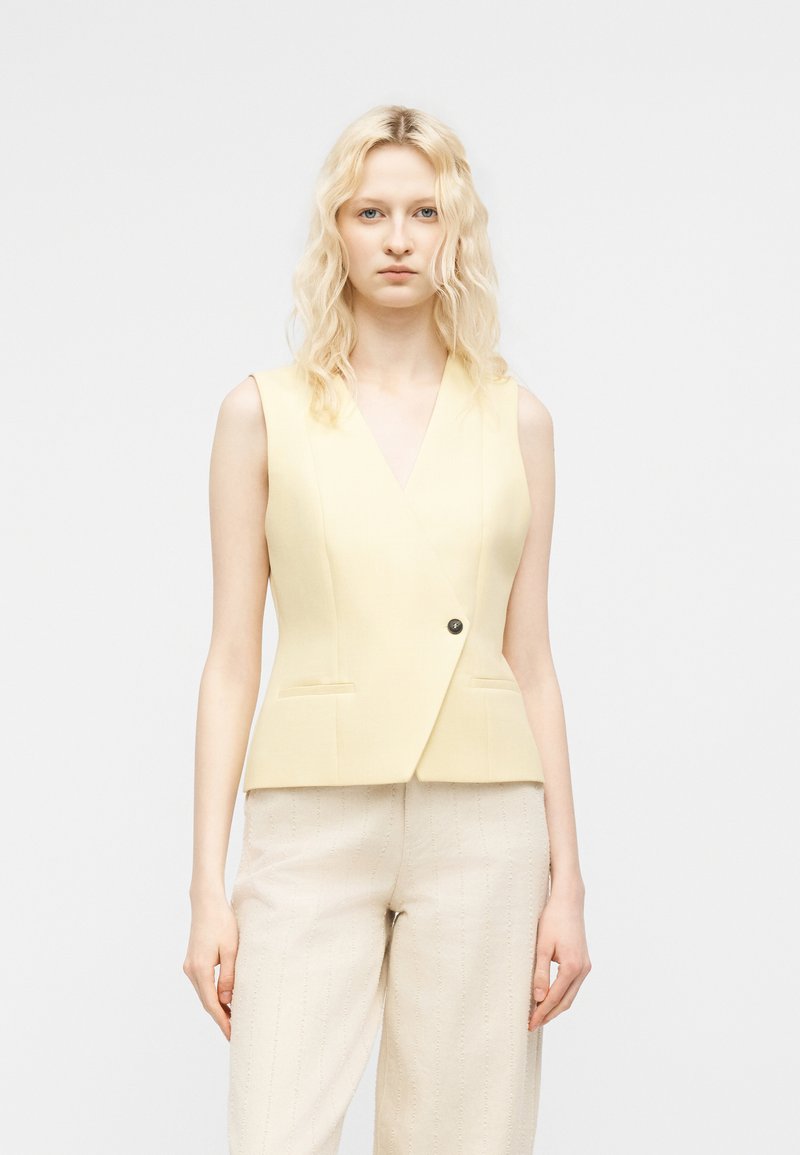 Γυναίκα με κυματιστά ξανθά μαλλιά φορά αμάνικο ανοιχτό κίτρινο γιλέκο με έναν κουμπί και κρεμ υφασμάτινο παντελόνι, με φόντο ένα απλό σκηνικό.