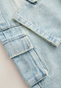 Svetlomodrá denim látka s textúrovaným povrchom, ktorá má manžetové rukávy a surové okraje. Obsahuje viditeľné švy.