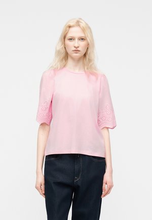 Blondynka w jasnoróżowej bluzce z krótkim rękawem z ażurowymi zdobieniami i ciemnych dżinsach, stojąca na tle jednolitego białego tła.