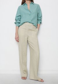 Camicia in lino turchese leggera con colletto e una tasca, abbinata a pantaloni in lino beige a gamba larga. Infradito color crema completano il look.