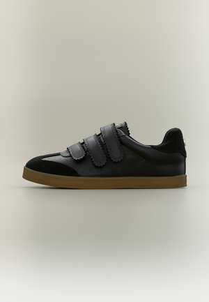 Baskets noires avec trois bandes velcro, détails perforés, embout en daim noir et semelle en caoutchouc marron sur fond neutre.