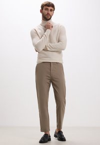 Mann in beigefarbenem Rollkragenpullover, braunen Slim-Fit-Hosen und schwarzen Quasten-Lederslippern, der mit einer Hand in der Nähe seines Kinns steht.