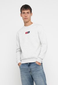Lichtgrijze katoenen sweatshirt met een ronde halslijn. Voorzien van een klein blauw en rood logo op de borst. Gecombineerd met blauwe denim jeans.