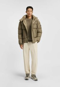 Puffjacka i olivgrön med hög krage och huva, med dragkedjestängning och elastiska ärmslut. Tillsammans med beige byxor och sneakers.