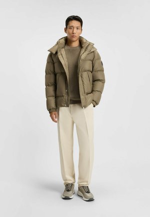 Giacca a vento verde oliva con colletto alto e cappuccio, dotata di chiusura a zip e polsini elasticizzati. Abbinata a pantaloni beige e sneakers.
