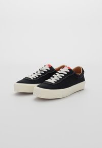 Svarta kanvas-sneakers med vita snören, rund tå och en kontrasterande vit gummisula. Röd logotypdetalj på tungan.
