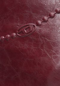 Dieprode leren oppervlak met een gegraveerde ketting van ronde kralen en een ovaal embleem met geometrische vormen in het midden.