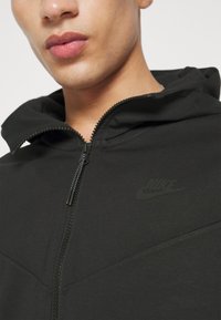 Czarna bluza z kapturem Nike na zamek błyskawiczny, wykonana z teksturowanej tkaniny, z regulowanym kapturem i subtelnym logo na klatce piersiowej, zaprojektowana w wąskim kroju.