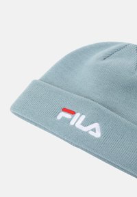 Bonnet tricoté bleu clair avec un revers, présentant un logo "FILA" brodé en blanc et un accent rouge à l'avant. Texture douce.
