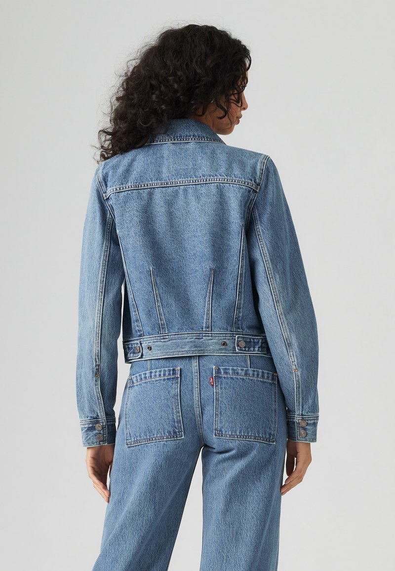 Chaqueta de mezclilla en azul claro con mangas largas, puños abotonados y bolsillos traseros. Presenta costuras clásicas y un diseño entallado en la cintura.