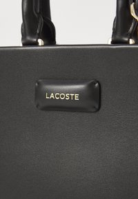Sac à main en cuir noir avec une surface texturée, deux poignées supérieures et une étiquette en cuir rectangulaire portant le logo doré "LACOSTE".