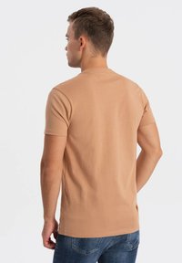 Herre kortærmet t-shirt i lys tan stof med en glat tekstur og rund hals, båret med blå jeans, set fra ryggen.