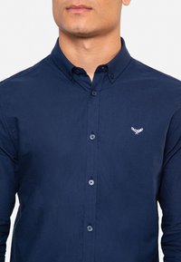 Camisa de botones azul marino de algodón, con cuello, mangas largas y un pequeño logotipo bordado en el pecho.