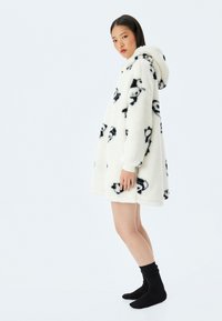 Abrigo capucha blanco esponjoso con estampados de cara de panda negra, mangas largas y una silueta relajada, combinado con calcetines negros.