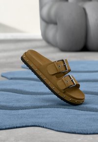 Καφέ σουέτ σανδάλια slides με δύο ρυθμιζόμενους ιμάντες και ασημένιες αγκράφες, με διακοσμητικές καρφίτσες κατά μήκος της σόλας. Υφή από καουτσούκ στη σόλα.