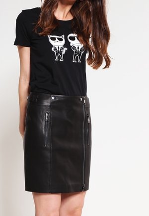 Frau trägt ein schwarzes T-Shirt mit Cartoon-Katzen mit Sonnenbrillen und einen schwarzen Lederrock mit vertikalen Reißverschlusstaschen und Frontreißverschluss.