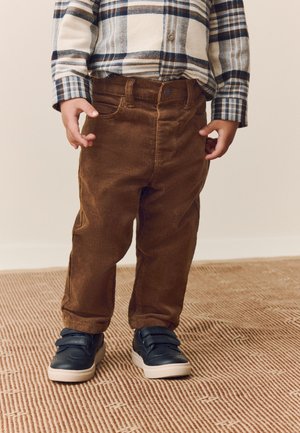 Kind draagt bruine ribfluwelen broek, een blauw-wit geruit overhemd en zwarte sneakers met witte zolen, staand op een gestructureerd tapijt.