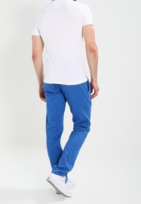 Bílé upnuté tričko, modré kalhoty a bílé tenisky. Kalhoty mají hladkou texturu a zúžený střih. Pohled ze zad.