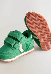 Zielone buty sportowe z rzepami, wykończone miękkim materiałem, z kontrastowymi białymi akcentami i gumową podeszwą z wzorem w jodełkę.