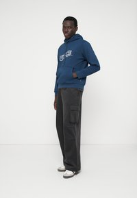 Tumši zils kapuci krekls ar grafikas dizainu, apvienots ar pelēkām kravas biksēm ar sānu kabatām. Modelis nēsā baltas un pelēkas tenisa kurpes.