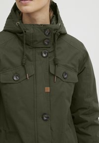 Veste vert olive en tissu durable, dotée d'une capuche, de plusieurs poches à boutons et de boutons décoratifs à l'avant.