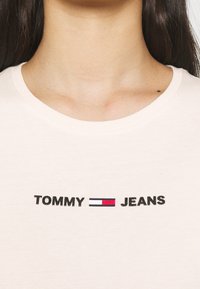 Camiseta de color beige claro con texto negro "TOMMY" y "JEANS" acompañado de un logotipo rectangular en rojo, blanco y azul en el centro. Diseño de cuello redondo, fabricada en algodón.