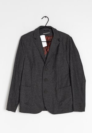 Blazer en chevrons gris foncé avec trois boutons, grandes poches avant et doublure intérieure à motif rouge, suspendu sur un cintre noir.