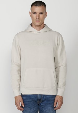 Haut à capuche beige clair en tissu doux, avec une poche frontale, un capuchon réglable, et un logo discret en relief sur la poitrine.