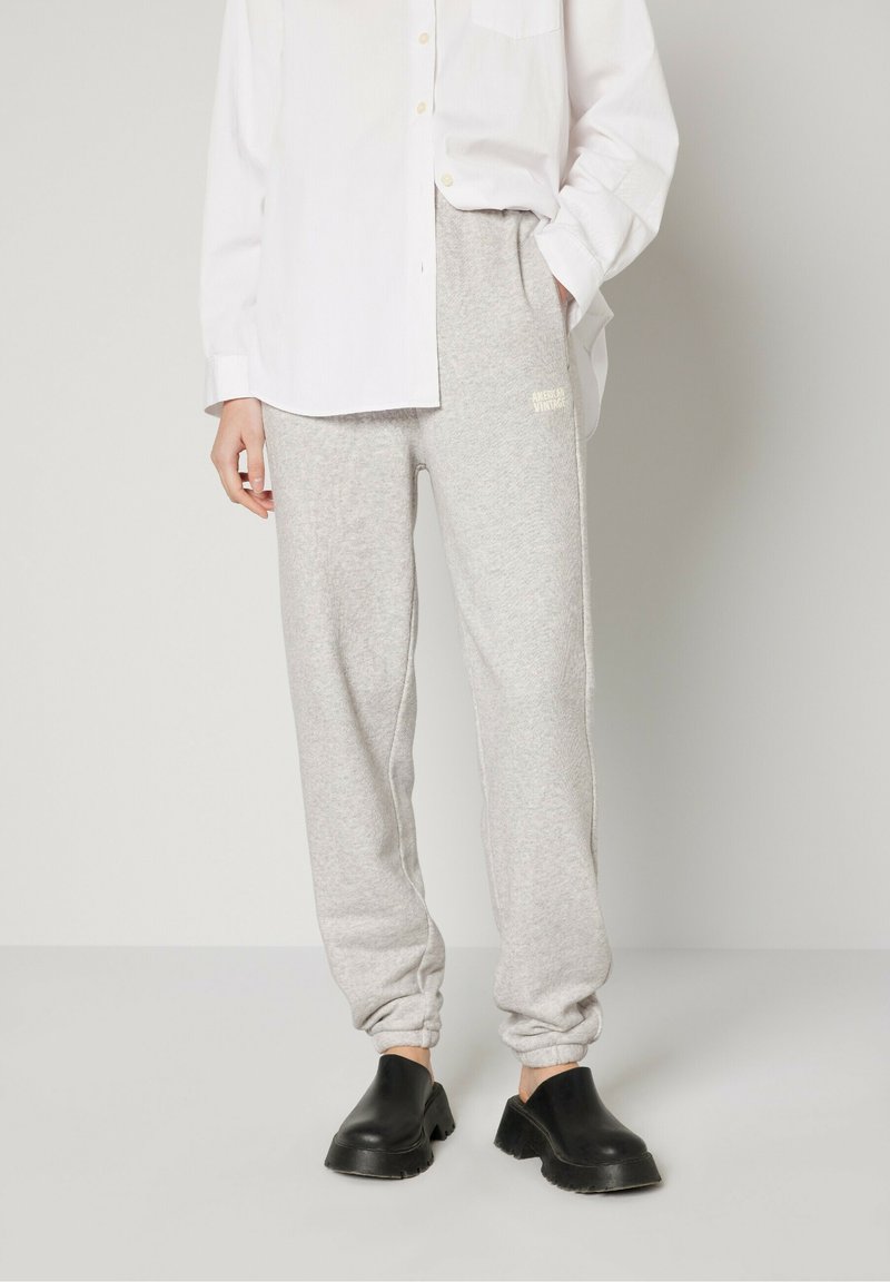 Pantalons de survêtement gris en tissu doux, avec des poignets élastiques et un logo vintage imprimé sur le côté gauche, associés à des sabots noirs.