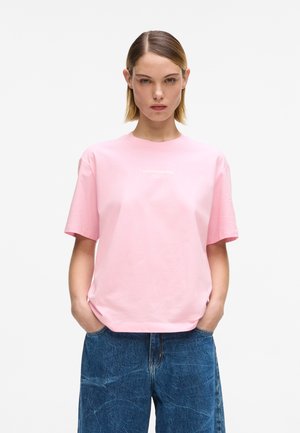 Frau mit glatten blonden Haaren trägt ein lockeres rosa T-Shirt und blaue Jeans aus Denim, steht mit den Händen in den Taschen vor einem weißen Hintergrund.