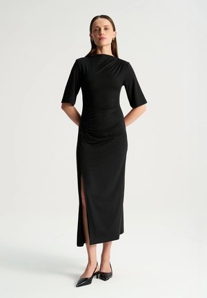 Kobieta stoi z rękami za plecami, ubrana w czarną sukienkę midi z rozcięciem po bokach i czarne szpiczaste szpilki na jednolitym jasnym tle.