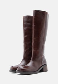 Barna bőr, térdig érő csizmák, lekerekített orral és vastag sarkú kivitelben. Simább tapintású, minimális varrási részletekkel, tartósságra tervezve.