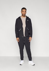 Musta hupullinen takki valkoisilla yksityiskohdilla, yhdistetty beigeen t-paitaan ja mustiin housuihin. Malli käyttää valkoisia urheilukenkiä.