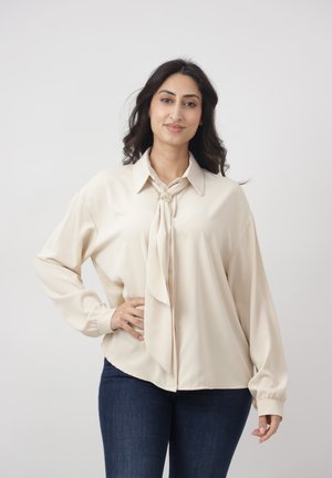 Kobieta o ciemnych, falujących włosach, ubrana w kremową bluzkę z ozdobną kokardą i ciemnoniebieskie jeansy, uśmiechająca się z jedną ręką na biodrze.