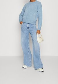 Hellblauer, flauschiger Strickpullover mit Rundhalsausschnitt, kombiniert mit weit geschnittenen, hellen Jeans und mehrfarbigen Sportschuhen. Hält eine kleine weiße Tasche.