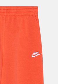 Oranžā kokvilnas bikses ar elastīgu jostasvietu. Apakšējā kreisajā kājā ir balts Nike logotips un mīksta, gluda tekstūra.