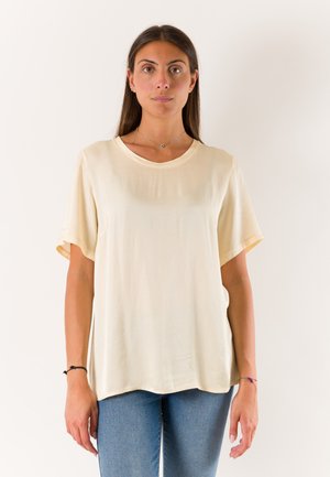 Donna con capelli lunghi e scuri che indossa una maglietta beige larga a maniche corte e jeans blu, in piedi davanti a uno sfondo chiaro e semplice.