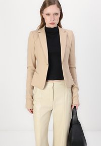 Bežový priliehavý blazer so strihom na mieru, dlhými rukávmi a trošku štruktúrovanými plecami. Skombinovaný s krémovými koženými nohavicami a čiernou taškou.