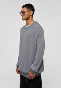 Grijze ribbeltrui met lange mouwen, ronde halslijn en een relaxte pasvorm. Draag met zwarte broek en accessorize met ronde zonnebril.