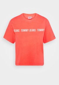 Korálové tričko s krátkým rukávem vyrobené z měkké bavlny, s výrazným bílým nápisem "TOMMY JEANS" na přední straně. Kulatý výstřih, uvolněný střih.