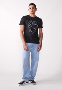 Zwarte T-shirt met witte leeuwgrafiek, van katoen, korte mouwen, gecombineerd met lichtblauwe jeans en zwarte sneakers met witte strepen.