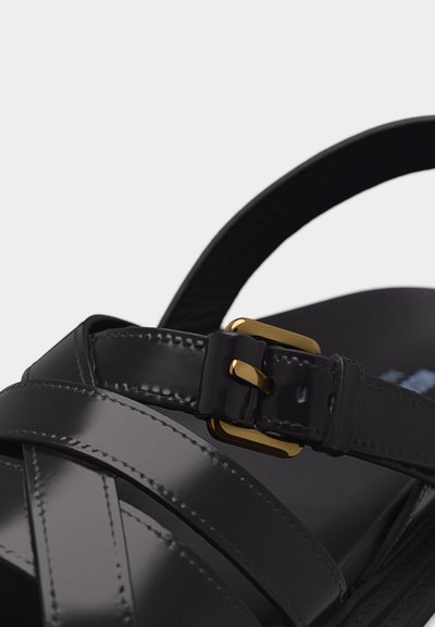 Μαύρο δερμάτινο σανδάλι με λουριά που διασταυρώνονται και αγκράφα σε χρυσό τόνο στη λουρίδα του αστραγάλου.