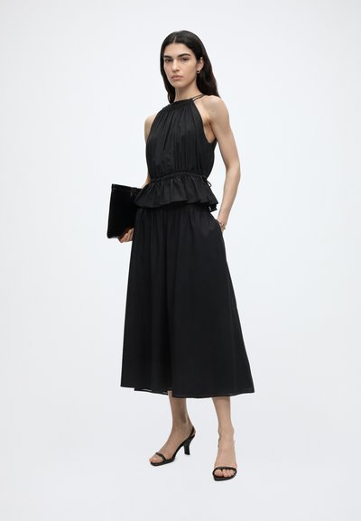 Γυναίκα με μακριά σκουρόχρωμα μαλλιά που φοράει ένα μαύρο αμάνικο τοπ halter με πεπλούμ και φαρδιά midi φούστα, κρατώντας ένα μαύρο clutch και φορώντας μαύρα τακούνια.